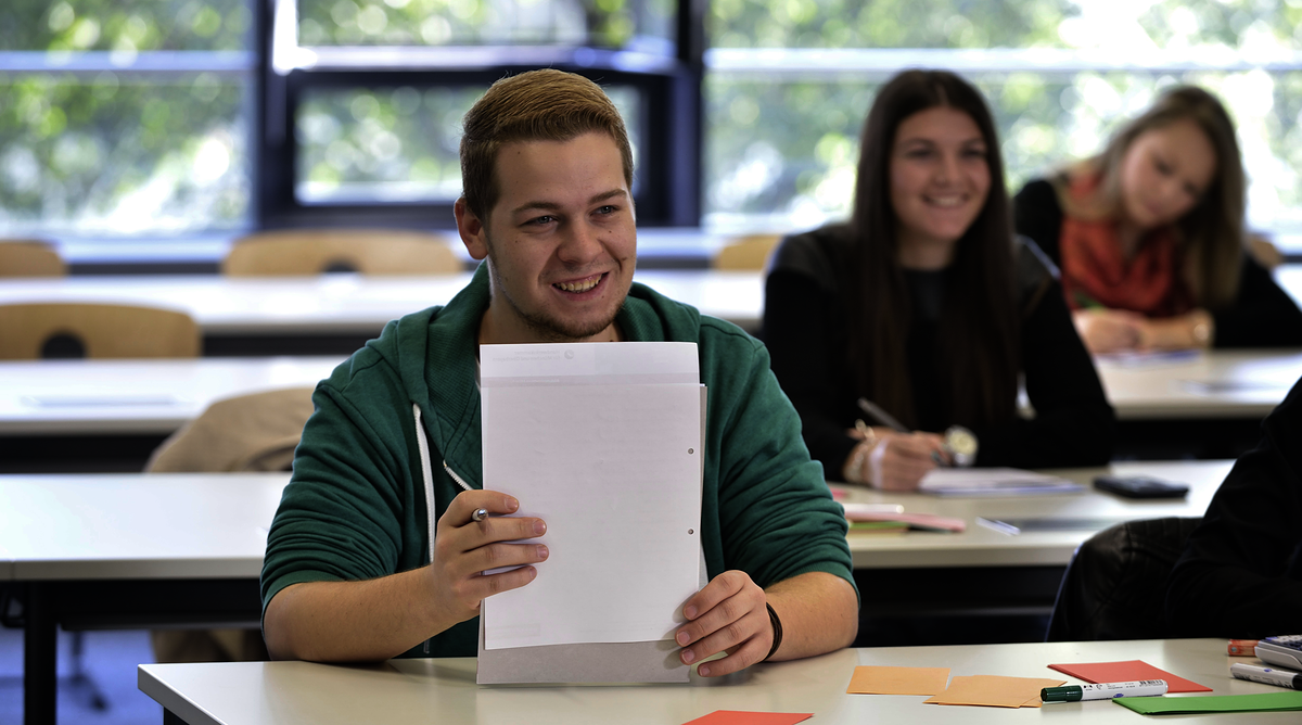 Prüfungswissen für Auszubildende Unterricht, Blick in die Klasse, lächelnder Azubi, Lehrling, hält Papier auf dem Tisch, Rund_um_die_Prüfung_15612_argum