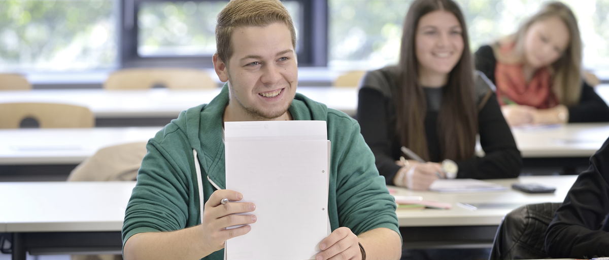 Prüfungswissen für Auszubildende Unterricht, Blick in die Klasse, lächelnder Azubi, Lehrling, hält Papier auf dem Tisch, Rund_um_die_Prüfung_15612_argum