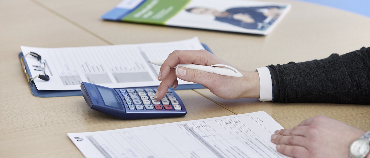 Nahaufnahme von Hand, Taschenrechner, Tisch mit Tabellen und Broschüre, Finanzierung_FH54261_argum