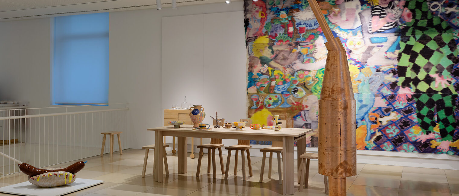 Impressionen mit Hotdog Foto mit großem buntem Gemälde, davor ein Hotdog aus Keramik, und eine große Skulptur aus Kupfer. Dazwischen ein eingedeckter Tisch.