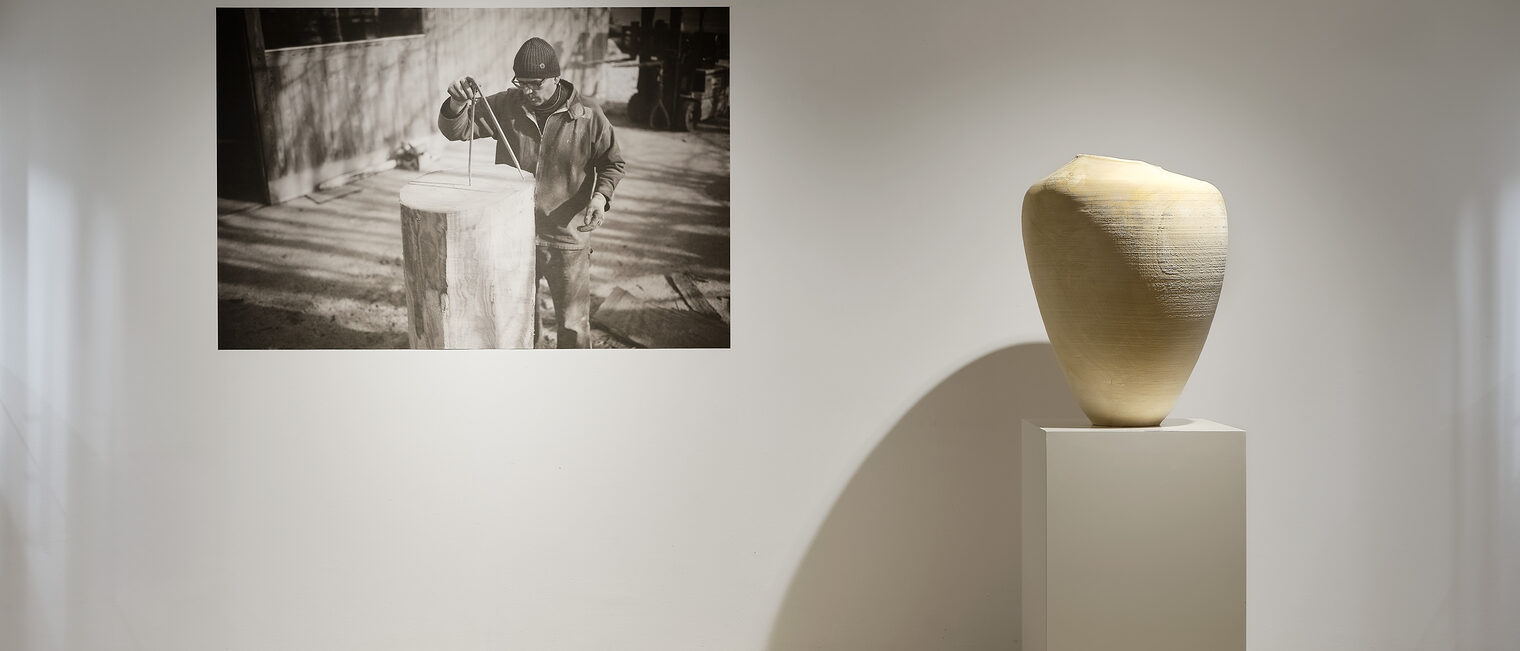 Eine große Schwarz-Weiß-Fotografie von Ernst Gamperl bei der Arbeit hängt neben einer hellen Holzskulptur, die bei der Galerie Handwerk ausgestellt ist.