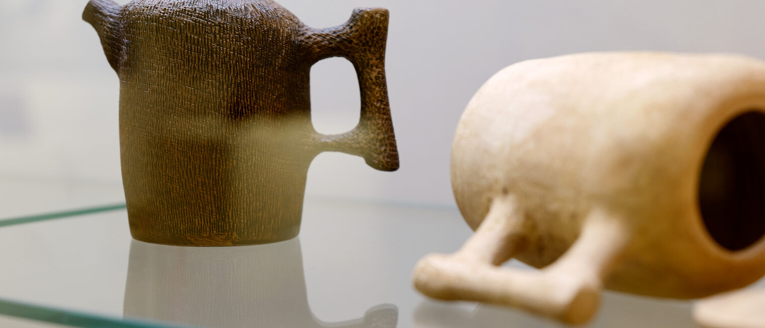 Eine dunkle Holz-Teekanne steht, eine helle Holz-Teekanne auf der Glasablage einer Vitrine. Sie spiegeln sich im Glas. Handwerkskunst von Ernst Gamperl
