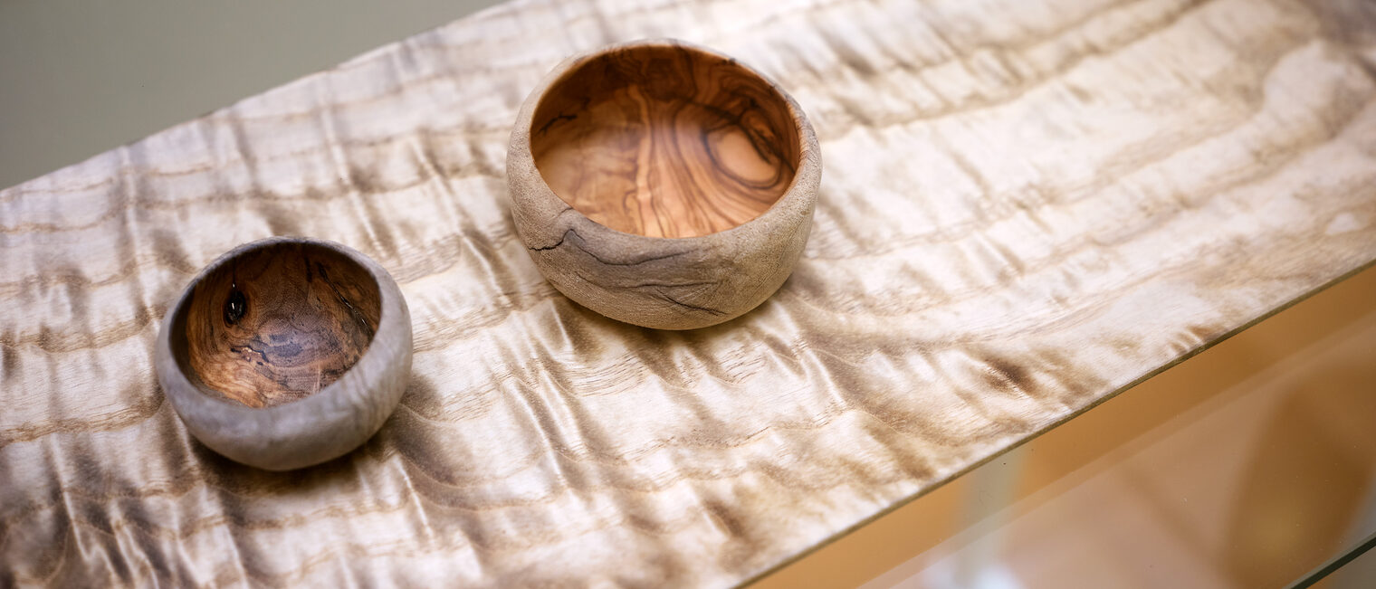 Zwei unterschiedlich große Holzschalen auf einem Holztisch, dessen intensive, wellenförmige Maserung plastisch wirkt.