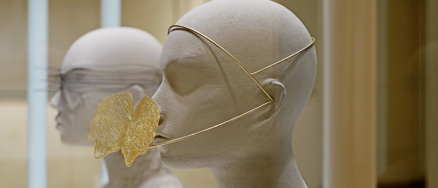 Zwei minimalistische graue Büsten in der Vitrine, mit abstraktem Schmuck aus Schnüren und filigranem Goldobjekt an der Nase.