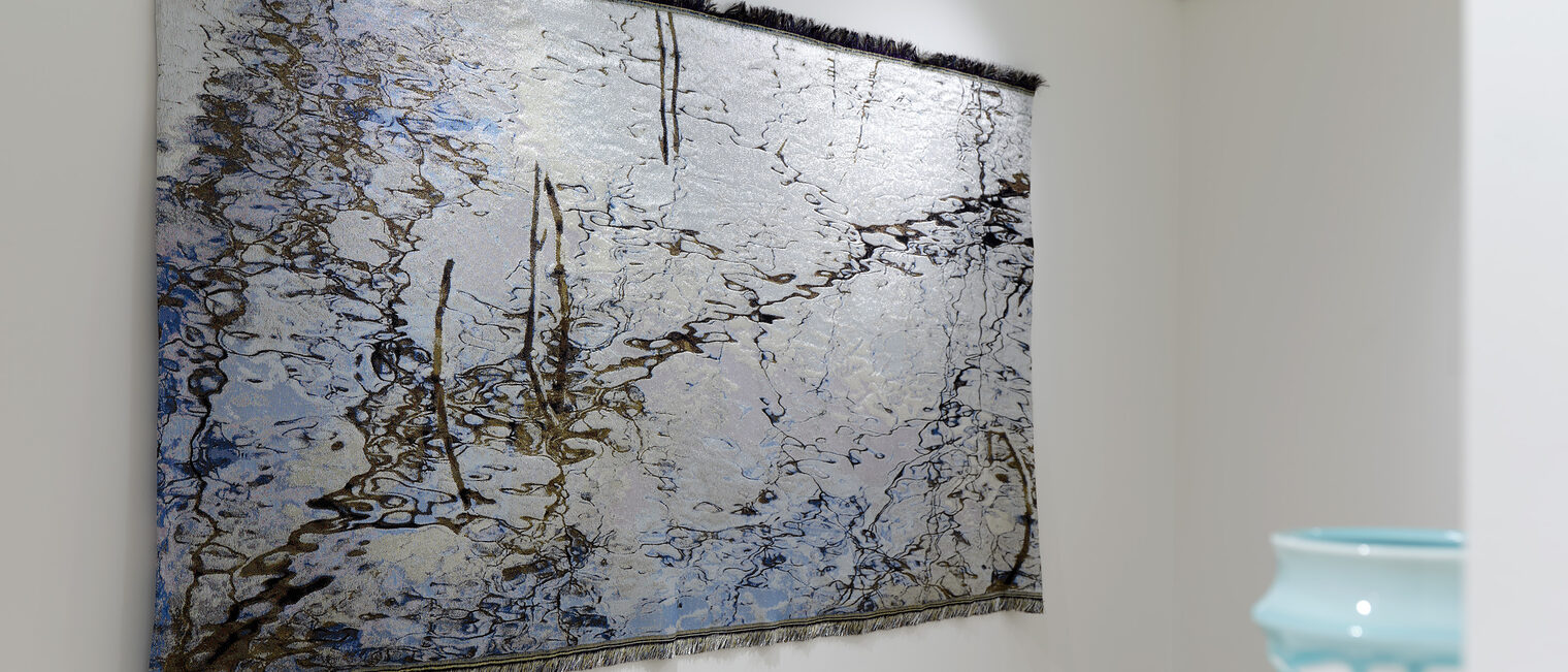 Ein fotorealistischer Wandteppich in zarten Farben, in dem sich der Himmel und Äste spiegeln - Ausstellung Galerie Handwerk