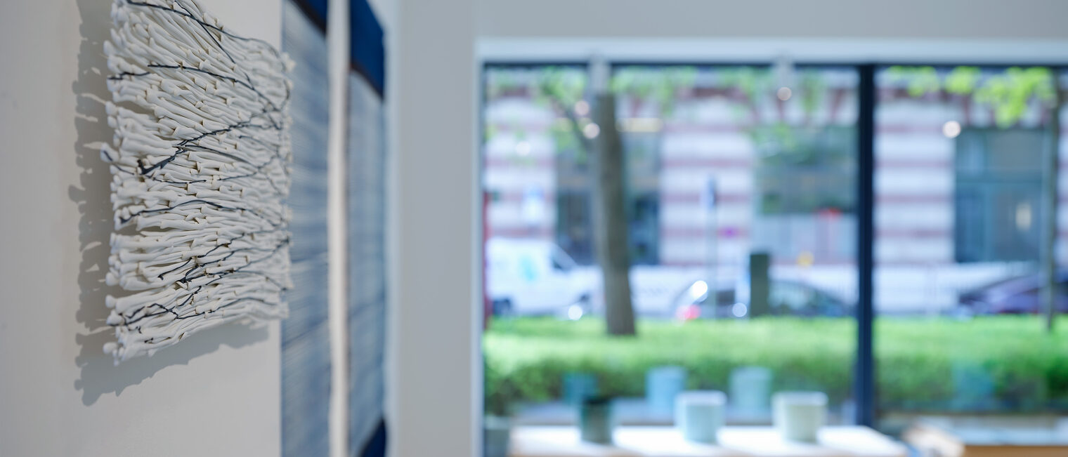Blick durch die Galerie nach draußen, an einem quadratischen Wandobjekt vorbei, das durch seine Struktur an Gischt erinnert.