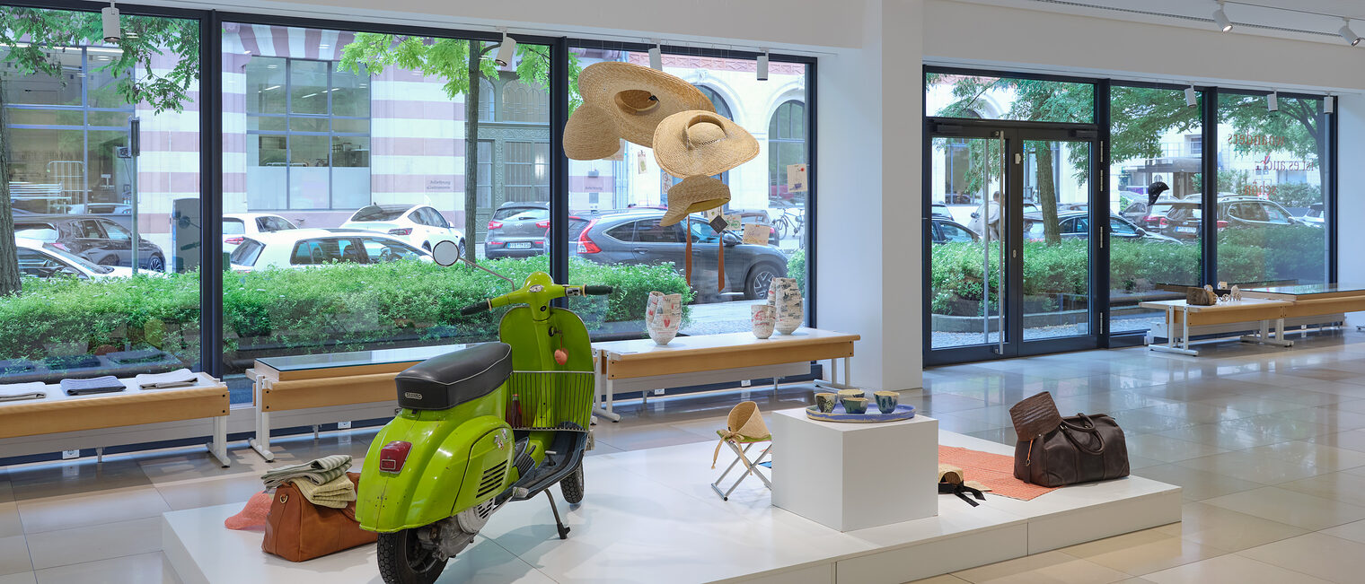 Blick aus der Galerie auf die Straße. Ensemble an Utensilien: Apfelgrüne Vespa, Reisetasche, Campingsitz, hängende Sonnenhüte