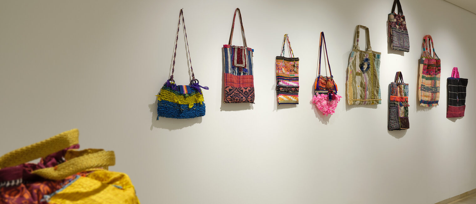Knallbunte Stofftaschen an der Wand, die unter anderem an Inka-Kunst und erinnern. Im Vordergrund eine bunte Reisetasche