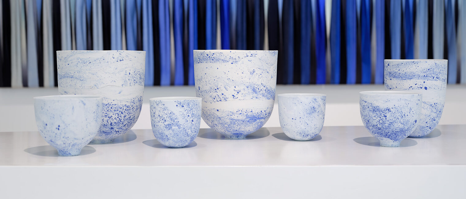 7 verschieden große Keramikbecher in zartblau-weiß marmoriert, dahinter transparente Stoffstreifen in mehreren Blautönen