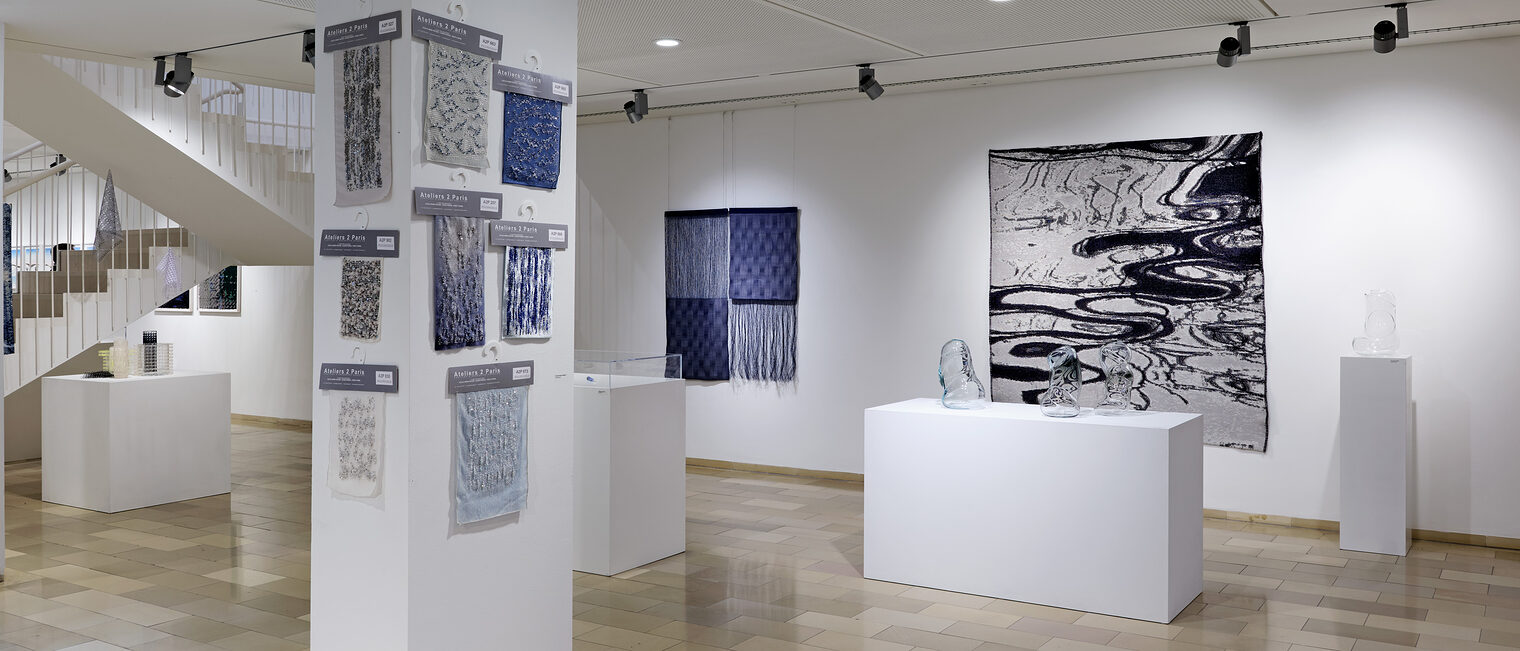 8 blaue Stoffmuster an der Säule. An der Wand ein großes Schwarz-Weiß-Tuch, das eine Wasseroberfläche fotorealistisch zeigt.