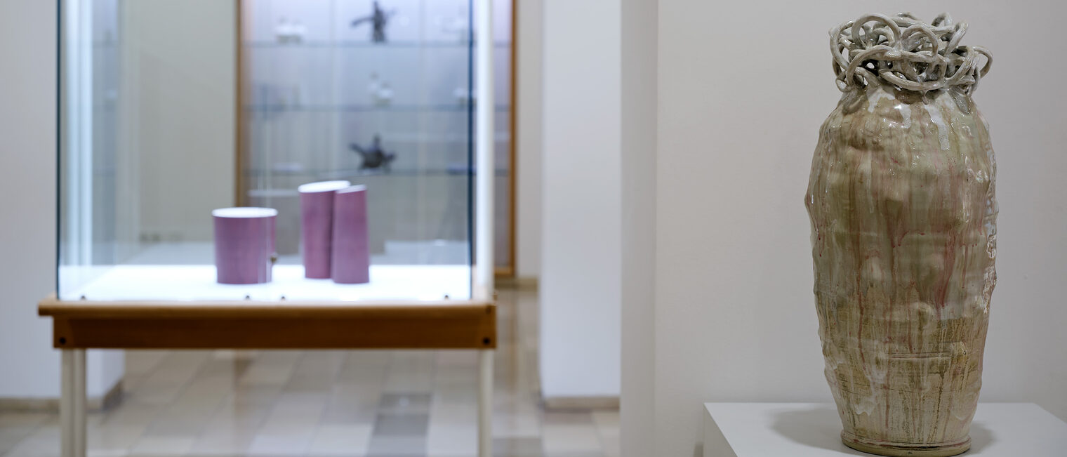 Große graubraune Vase, auf deren Öffnung sich Schlingen aus Keramik winden. Dahinter Vitrine mit drei violetten geraden Vasen